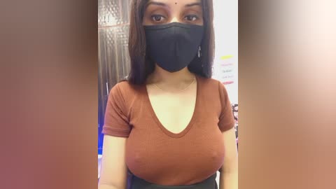 Video of a young woman with long dark hair, wearing a black face mask and a rust-colored ribbed short-sleeve top, standing indoors near a glass door.