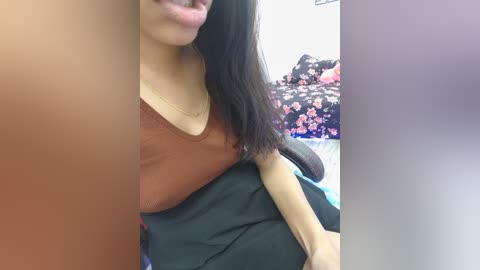 A close-up video of a young Asian woman with long black hair, wearing a brown tank top and sitting on a bed with a floral-patterned bedspread.