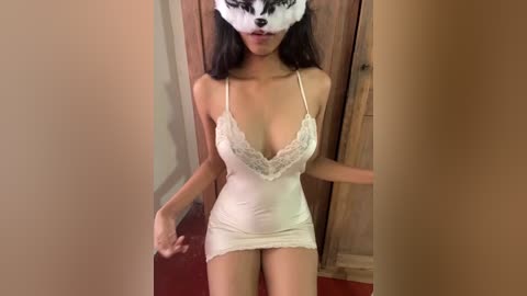 Video of an Asian woman in a white, lace-trimmed, low-cut nightgown, standing in a hallway with wooden doors, wearing a fox mask.