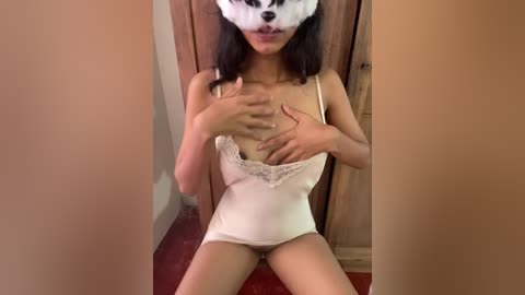 A video of a young Asian woman with long black hair and light skin, wearing a white lace-trimmed camisole, sitting on a red carpet. She has a white mask with a black dog face covering her face.