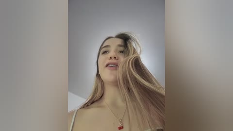 Video of a young woman with long, straight blonde hair, fair skin, and a slender physique. She wears a white tank top and a red necklace, with a slightly blurred background.
