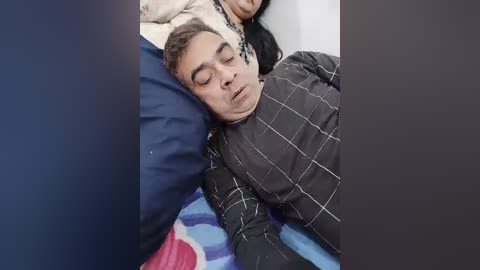 Video of a sleeping man in a dark patterned shirt on a blue bed with a pink blanket, partially obscured by a dark object.