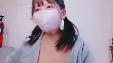 Video of an Asian woman with long black hair, wearing a light blue cardigan, white surgical mask, and large white earrings, in a cluttered room with a red dress hanging on the left and a white shelf on the right.