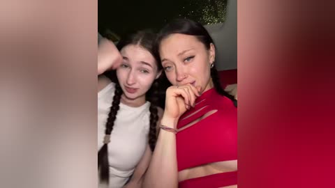 Video of two young women with fair skin, one with braided hair and a white top, the other with dark hair and a red top, posing closely together, smiling.