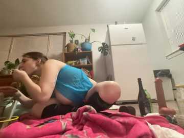 Video of a curvy woman with fair skin, in a blue tank top and black shorts, kneeling on a bed with a pink blanket, surrounded by scattered items in a cluttered bedroom.