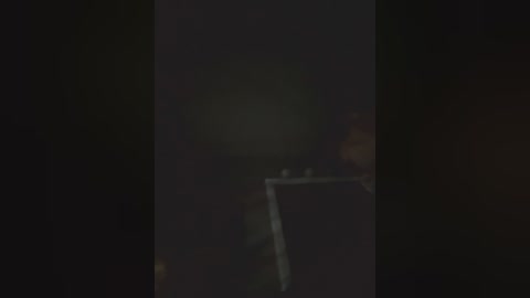 A dimly lit video showing a man with a beard and a dark shirt, partially obscured by shadows, standing next to a railing. The background is indistinct and dark.