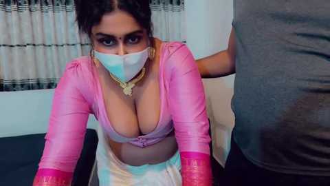A video of a dark-skinned woman with large breasts, wearing a pink crop top and white skirt, kneeling on a couch, masked, and restrained by a man.