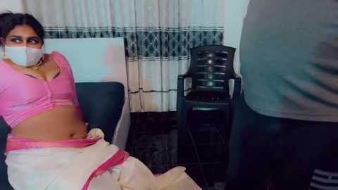 Video of a young woman with medium skin tone, wearing a pink crop top, white mask, and pink saree, sitting on a blue sofa, in a room with white curtains, and a black chair.