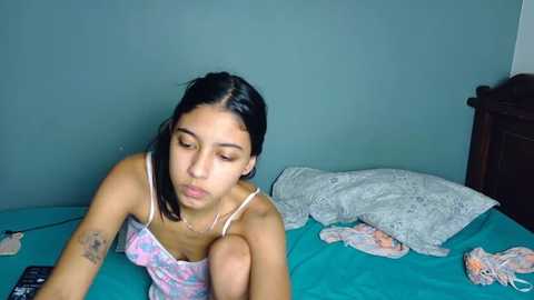 Video of a Latina woman with straight black hair, medium build, wearing a pink and white patterned camisole, lying on a turquoise bedspread in a teal room, surrounded by disheveled bedding.