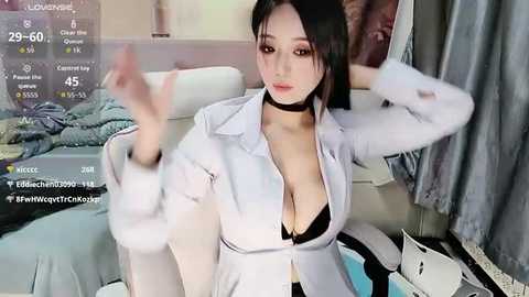 Video of an East Asian woman with long black hair, wearing a white lab coat, black choker, and revealing black bra, sitting on a bed, surrounded by messy clothes and a digital screen displaying weather and social media.