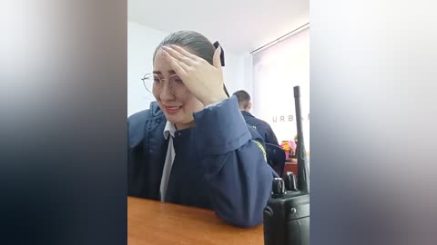 A video of a woman with fair skin, wearing glasses, a navy blue jacket, and a gray tie, covering her forehead with her right hand while smiling, seated at a wooden desk.