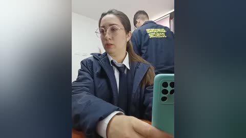 Video of a young Asian woman with light skin, wearing glasses and a dark navy jacket, seated at a table in a dimly lit room. Behind her, another person in a similar jacket is visible.