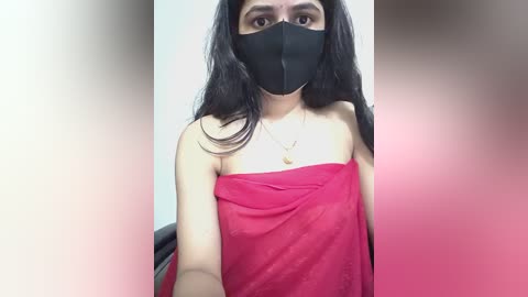 Video of a South Asian woman with long black hair, wearing a black face mask, a red cloth covering her shoulders, and a gold necklace. The background is blurred in a pink gradient.
