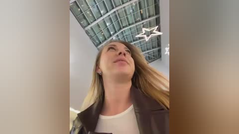 Video of a young woman with long blonde hair, wearing a black leather jacket, looking upward at a glass ceiling with star-shaped lights, in a modern, possibly airport setting.