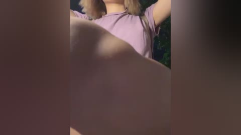 Video of a blonde woman in a light purple top, arms raised, with blurred, out-of-focus objects in the foreground.