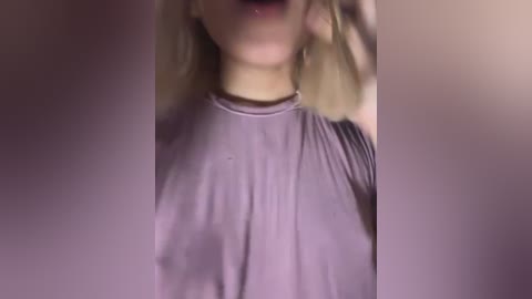 Video of a woman in a loose-fitting, light purple top, with her face partially obscured by blurred, darkened edges, suggesting a dimly lit, intimate setting.