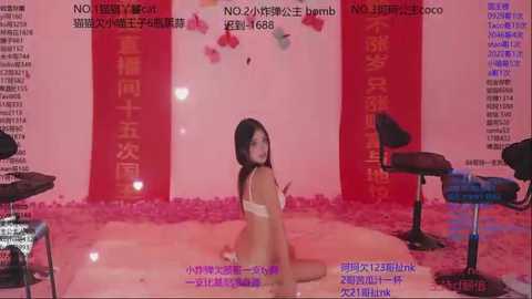 A video of an Asian woman in lingerie kneeling on a shiny pink floor, surrounded by red banners and stools, set in a red and pink room.