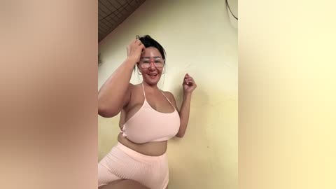 A video of a smiling, plus-sized woman with medium skin tone and short black hair, wearing glasses and a light pink sports bra and high-waisted shorts, posing against a yellow wall in a dimly lit room.