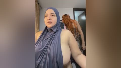 Video of a plus-size woman in a blue hijab, partially nude, with a large cat in a room with beige walls and a door.
