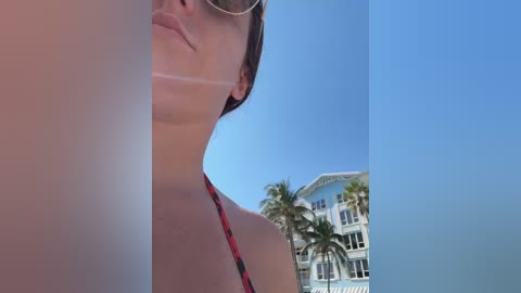 Video of a woman in a red bikini, with a tan line on her neck, standing next to a white house with palm trees.