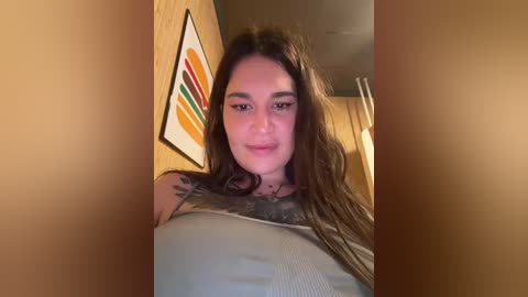 Video of a young woman with long, wavy brown hair, fair skin, and a tattoo on her chest, wearing a gray tank top. She stands in front of a wooden wall with a colorful abstract painting.