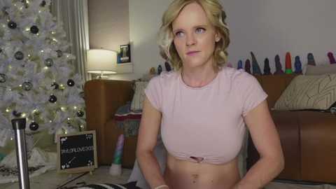 Video of a blonde woman with short, wavy hair wearing a pink crop top, sitting indoors. Christmas tree, toys, and a sign with \"BLOWJOB\" in the background.