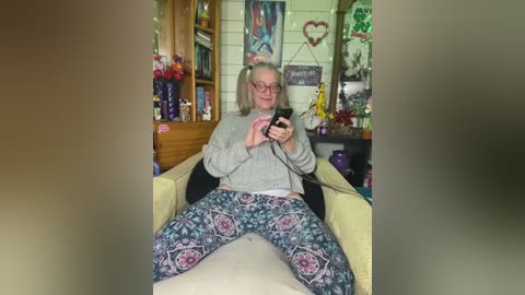 A video of an elderly woman with glasses, gray hair, and a gray sweater, sitting on a beige couch, wearing patterned pajama pants, holding a phone, in a cozy, cluttered living room with wooden bookshelves, decorative items, and colorful artwork.