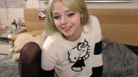 Video of a smiling young woman with long, straight, blonde hair, wearing a black-and-white Hello Kitty shirt, black leggings, and sitting on a bed with plush toys and a wooden headboard.