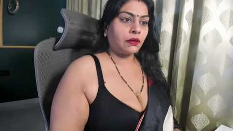 A video of a South Asian woman with long black hair, wearing a black bra and necklace, sitting in a gray chair with a beige cushion. The background includes dark green curtains and a floral-patterned curtain.