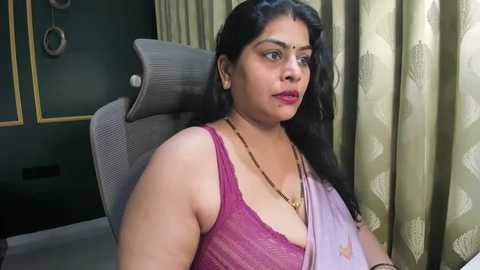 Video of a South Asian woman with medium skin tone, wearing a pink lace bralette, sitting on a grey office chair. She has long black hair, minimal makeup, and a serious expression.