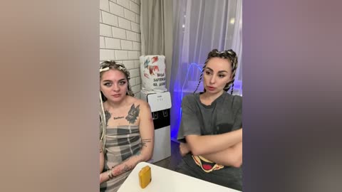 Video of two young women in a modern caf\u00e9. One wears a gray T-shirt, the other a plaid apron; both have tattoos. Background includes white brick wall, water cooler, and semi-transparent curtains.