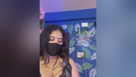 A video of a South Asian woman with medium skin tone, wearing a black mask, black dress, and colorful jewelry, standing in a room with blue walls adorned with tropical leaf stickers and green plants.