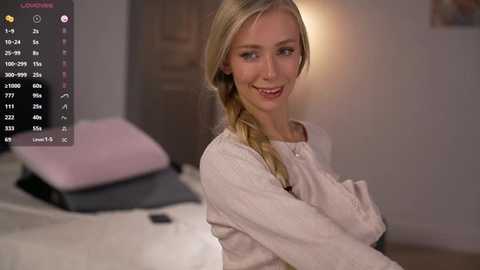 Video of a smiling, fair-skinned blonde woman in a beige sweater, seated on a bed with pink and white pillows in a softly lit bedroom.