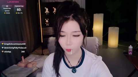 Video of an East Asian woman with black hair, wearing a white top, with semen on her face, in a dimly lit room with candles and a bed.