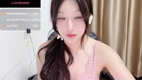 Video of an East Asian woman with long black hair, wearing a pink lace bra and white headphones, sitting in front of a beige curtain, looking tired.