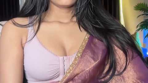 Video of a South Asian woman with long, straight black hair and medium skin tone, wearing a purple blouse with a plunging neckline and a maroon sari with gold embroidery, indoors with blurred background.