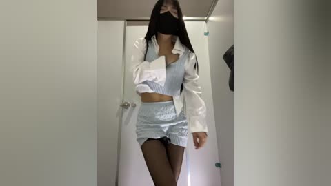 A video of an Asian woman in a white lab coat, black face mask, and light blue shorts, walking down a corridor with a white door in the background.