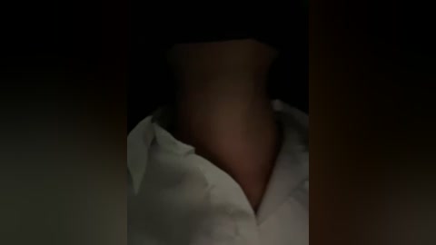Video of a person's neck and shoulders, partially obscured by a dark, blurry background. They wear a white collared shirt with buttons. The lighting is dim, creating a moody atmosphere.
