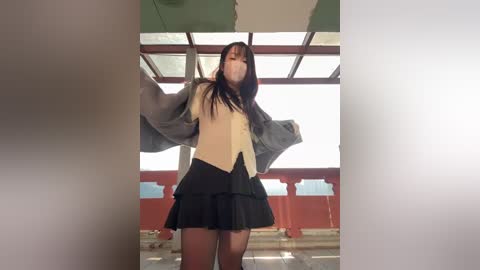 A video of an Asian woman with long black hair, wearing a beige vest over a grey sweater, black skirt, and face mask, standing in a dimly lit, red-roofed building with large windows.