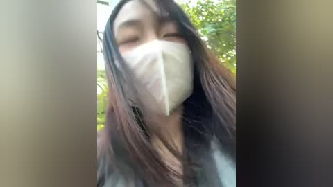 Video of an Asian woman with long, straight black hair, wearing a white surgical mask, standing outdoors with blurred greenery in the background.
