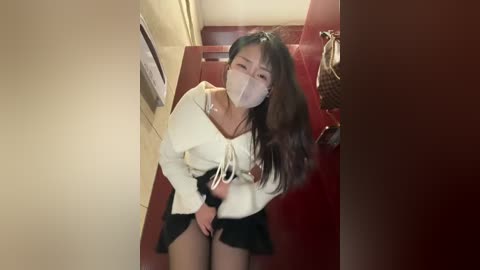 A video of an Asian woman in a white top and black skirt, wearing a face mask, bending over in a dimly lit hallway with a red door and wicker basket in the background.