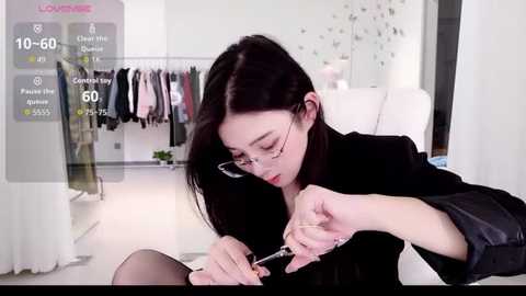 Video of an East Asian woman with fair skin and long black hair, wearing glasses, adjusting a black dress in a modern, well-lit dressing room with hanging clothes.