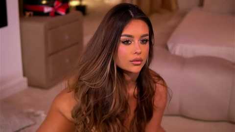 Video of a topless, light-skinned woman with long, wavy brown hair, wearing dramatic makeup, sitting in a dimly lit bedroom with beige furniture and a bed.