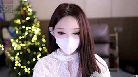 A video of an East Asian woman with long, straight brown hair, wearing a white lace dress and a face mask, standing in front of a Christmas tree with glowing lights.