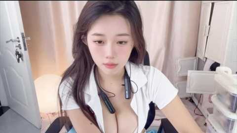 Video of an Asian woman with long black hair, wearing a white shirt, exposing ample cleavage, headphones around her neck, seated in a cluttered room with beige curtains and white furniture.