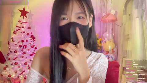 A video of an Asian woman with long black hair, wearing a white lace top and black mask, posing with a finger to her lips in a festive room with a decorated Christmas tree and pink lights.