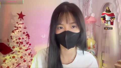 A video of an Asian woman with long black hair, wearing a black face mask and white top, standing in a festive room with a decorated Christmas tree and a \"Halloween\" sign.