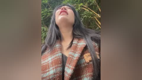 Video of a woman with long, straight black hair, wearing a brown plaid coat, standing outside against a background of green bamboo, her head tilted back, smiling.