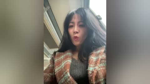 Video of an Asian woman with long, dark hair and a medium complexion, wearing a brown and white plaid sweater over a black top, making a kissing face indoors near a window.