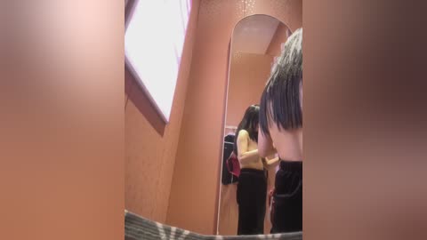 A video of a person with dark hair, wearing a black shirt, standing in a narrow, peach-painted hallway with a mirror and a white light fixture.